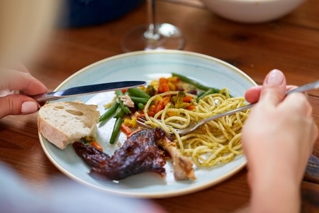 food and people concept - hands of woman with fork and knife eating pasta and roast chicken at wooden tableの写真素材