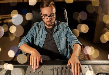music, technology, people and equipment concept - man at mixing console in sound recording studio...