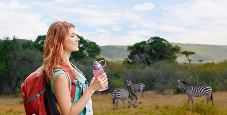 adventure, travel, tourism, hike and people concept - smiling young woman with backpack and bottle of water over zebras in african savannah backgroundの写真素材