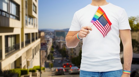 lgbt, same-sex relationships and homosexual concept - close up of man wearing gay pride rainbow awareness wristbands and holding american flag over san francisco city backgroundの写真素材