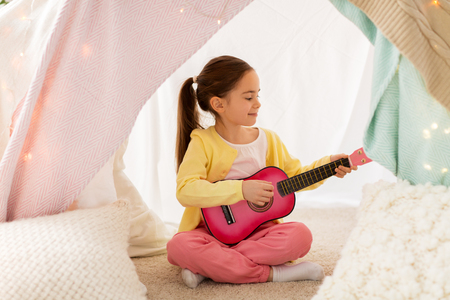 childhood and hygge concept - happy little girl playing toy guitar in kids tent at homeの写真素材