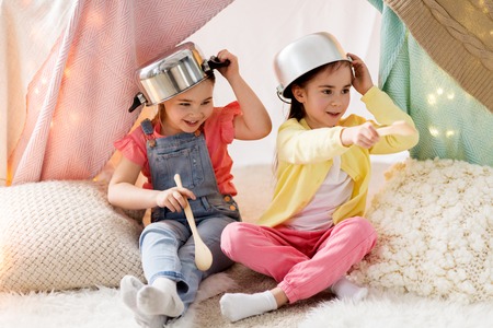 childhood and hygge concept - happy little girls with cooking pots playing in kids tent at homeの写真素材