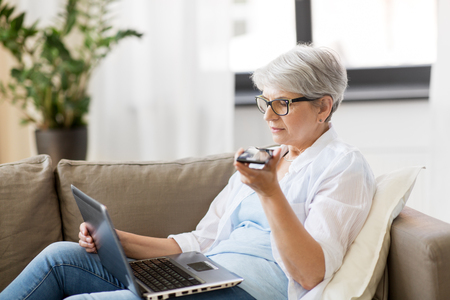 technology, old age and people concept - happy senior woman in glasses with laptop computer and smartphone using voice command recorder at homeの写真素材