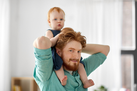 family, fatherhood and people concept - happy red haired father carrying his little baby daughter on neck at homeの写真素材