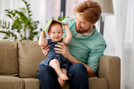family, fatherhood and people concept - happy red haired father and little baby daughter playing with book at homeの写真素材