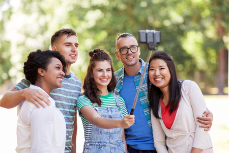 people, friendship and international concept - happy smiling young woman and group of happy friends taking picture by selfie stick in parkの写真素材
