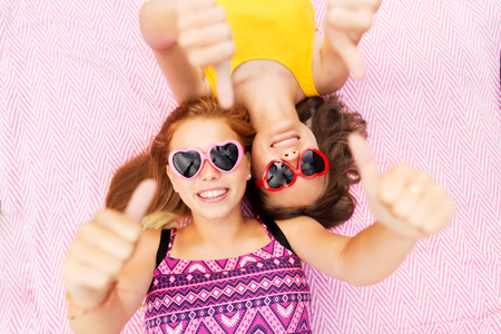 summer fashion, leisure and valentines day concept - smiling teenage girls in heart shaped sunglasses lying on picnic blanket and showing thumbs upの写真素材