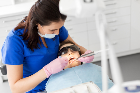 medicine, dentistry and healthcare concept - female dentist with mouth mirror and water gun checking for kid patient teeth at dental clinicの写真素材