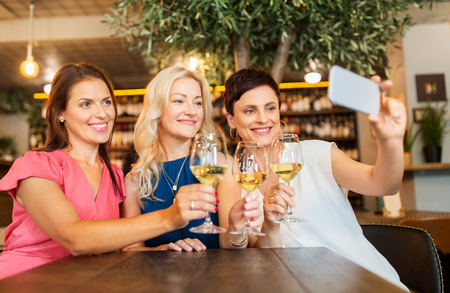 people, technology and lifestyle concept - women drinking wine and taking selfie by smartphone at bar or restaurantの写真素材