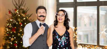 celebration, fun and holidays concept - happy couple posing with party props over christmas tree...