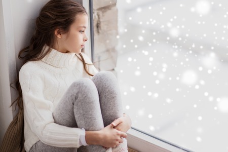 sad girl sitting on sill at home window in winterの写真素材