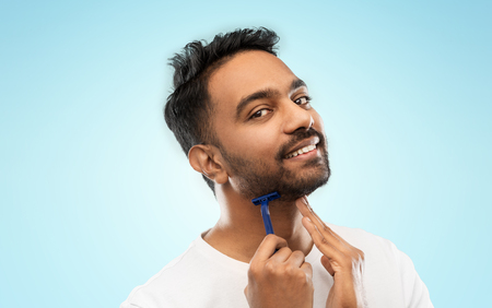 grooming and people concept - smiling indian man shaving beard with manual razor blade over blue backgroundの写真素材
