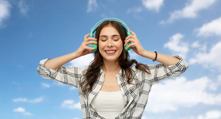 music, technology and people concept - happy young woman or teenage girl with headphones over blue sky and clouds backgroundの写真素材