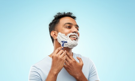 grooming and people concept - young indian man shaving beard with manual razor blade over blue backgroundの写真素材