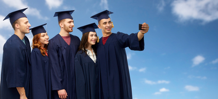education, graduation and people concept - group of happy graduate students in mortar boards and bachelor gowns taking selfie by smartphone over blue sky and clouds backgroundの写真素材