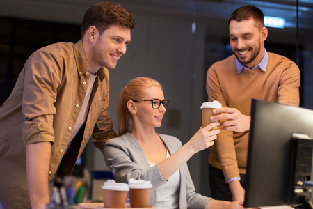 business, deadline and people concept - coworkers with computer and takeout coffee working late at night officeの写真素材