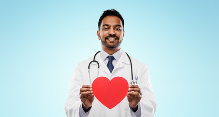 medicine, cardiology and healthcare concept - smiling indian male doctor or cardiologist in white coat with red heart shape and stethoscope over blue backgroundの写真素材