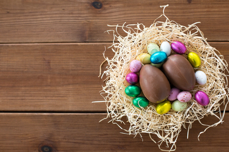 easter, confectionery and holidays concept - chocolate eggs and candies in straw nest on wooden backgroundの写真素材