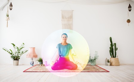 mindfulness, spirituality and healthy lifestyle concept - woman meditating in lotus pose at yoga studio over rainbow auraの写真素材