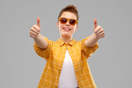 summer, eyewear and people concept - smiling red haired teenage girl in sunglasses and yellow checkered shirt showing thumbs up over grey backgroundの写真素材