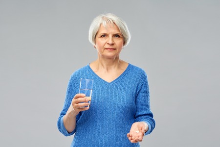 Medicine, health and old people concept - senior woman with glass of water and pills over grey backgroundの写真素材
