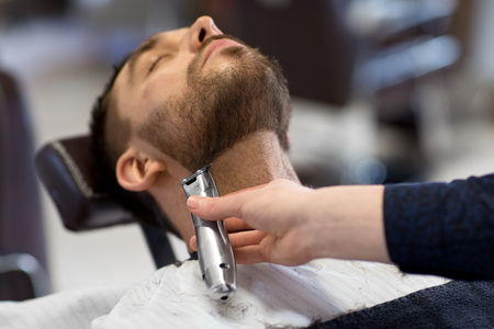 grooming and people concept - man and barber with trimmer or shaver cutting beard at barbershopの写真素材