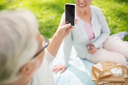 old age, leisure and technology concept - close up of happy senior couple having picnic and taking picture by smartphone at summer parkの写真素材