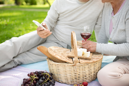 old age, holidays, leisure and people concept - close up of senior couple with smartphone, picnic basket and red wine sitting on blanket at summer parkの写真素材