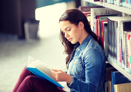 education, high school, university, learning and people concept - student girl reading book sitting on floor at libraryの写真素材