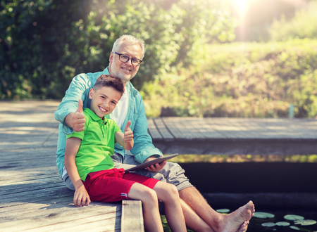 family, generation, technology, communication and people concept - grandfather and grandson with tablet pc computer sitting on river berthの写真素材