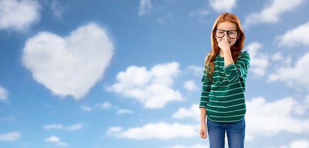 education, vision and valentine's day concept - cute smiling red haired student girl in glasses and green striped shirt over blue sky and heart shaped cloud clouds backgroundの写真素材