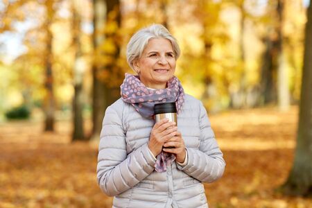Old woman with hot drink in tumbler at autumn park