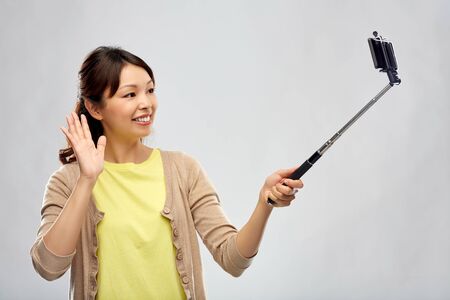 technology and people concept - happy asian woman taking picture by smartphone on selfie stick and waving hand over grey backgroundの写真素材