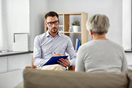 geriatric psychology, mental therapy and old age concept - psychologist with clipboard taking notes and listening to senior woman patient at psychotherapy sessionの写真素材