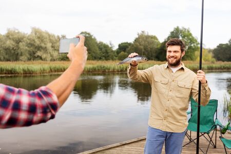 fishing, leisure and people concept - friend photographing fisherman with fish by smartphone at lakeの写真素材