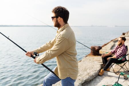 leisure and people concept - male friends with fishing rods on pier at sea