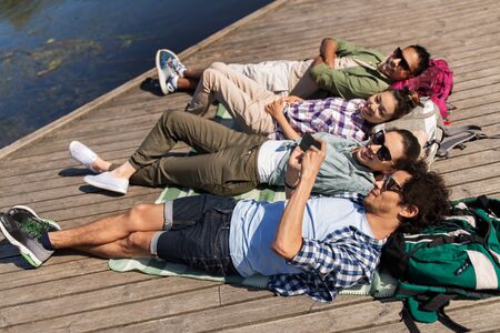 friends with smartphone lying on lake pier