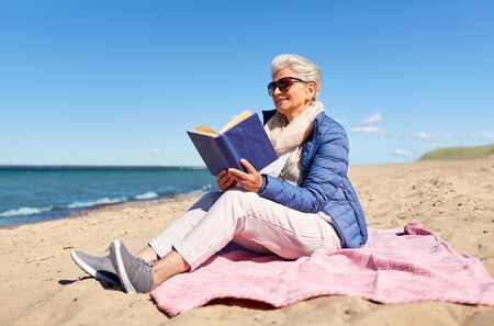 people and leisure concept - happy senior woman reading book on summer beach in estoniaの写真素材