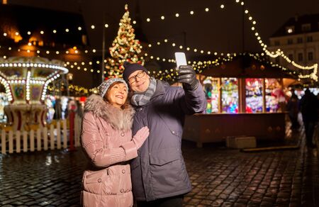 Happy senior couple taking selfie by smartphone at christmas market on town hall square