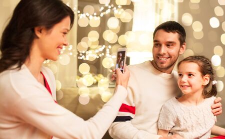 Happy mother, father and little daughter having christmas dinner and taking picture by smartphone at...