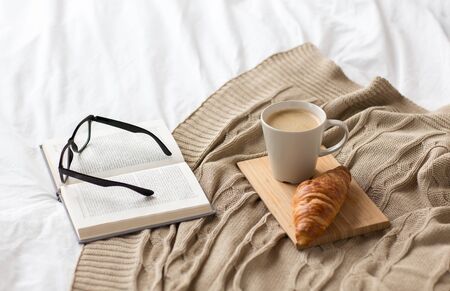 coffee, croissant, blanket and book on bed at home