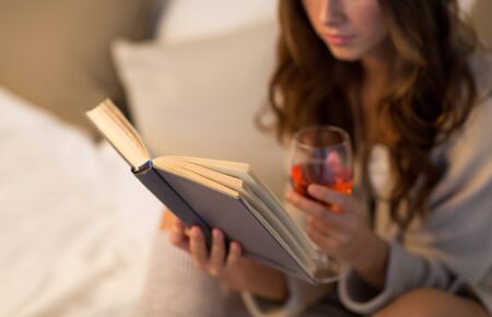 christmas, leisure and comfort concept - close up of young woman reading book and drinking rose wine from glass at homeの写真素材