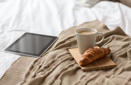 comfort, hygge and interior concept - tablet computer, cup of coffee and croissant on bed at cozy homeの写真素材
