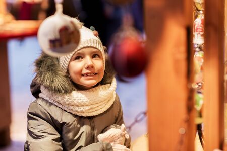 holidays, childhood and people concept - happy little girl at christmas market in winter eveningの写真素材