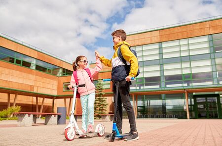 education, childhood and people concept - happy school children with backpacks and scooters making high five outdoorsの写真素材