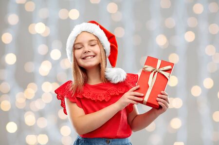 christmas, childhood and holidays concept - smiling pleased girl posing in santa helper hat with gift box over festive lights background