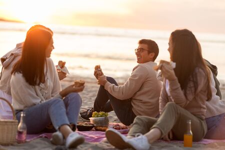 happy friends eating sandwiches at picnic on beachの写真素材