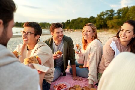 happy friends eating sandwiches at picnic on beach