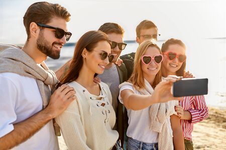 friendship, leisure and people concept - group of happy friends taking selfie by smartphone on beach in summerの写真素材