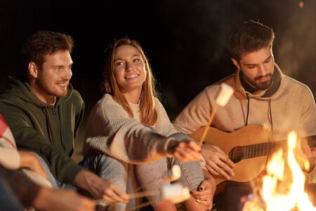 leisure and people concept - group of smiling friends sitting at camp fire, roasting marshmallow and...